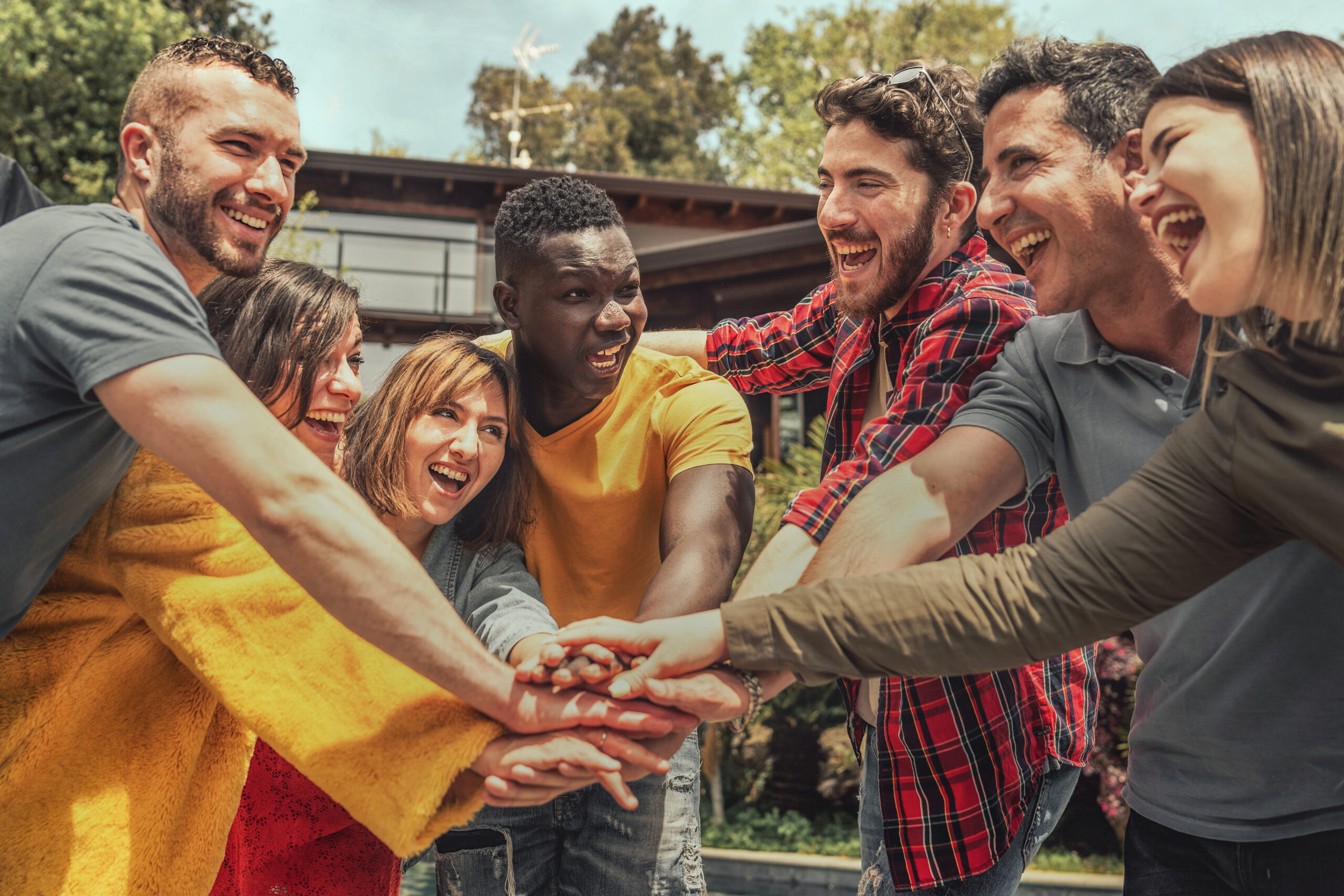 A multiracial group of young individuals forms a circle outdoors, placing their hands on top of one another's. They celebrate joyfully, looking into each other's eyes, embodying unity and friendship.